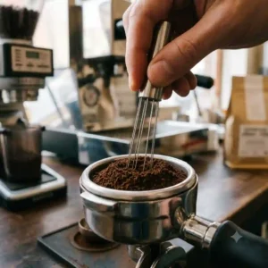 A barista uses a needle distribution tool to break clumps in ground coffee for espresso machine within a portafilter.